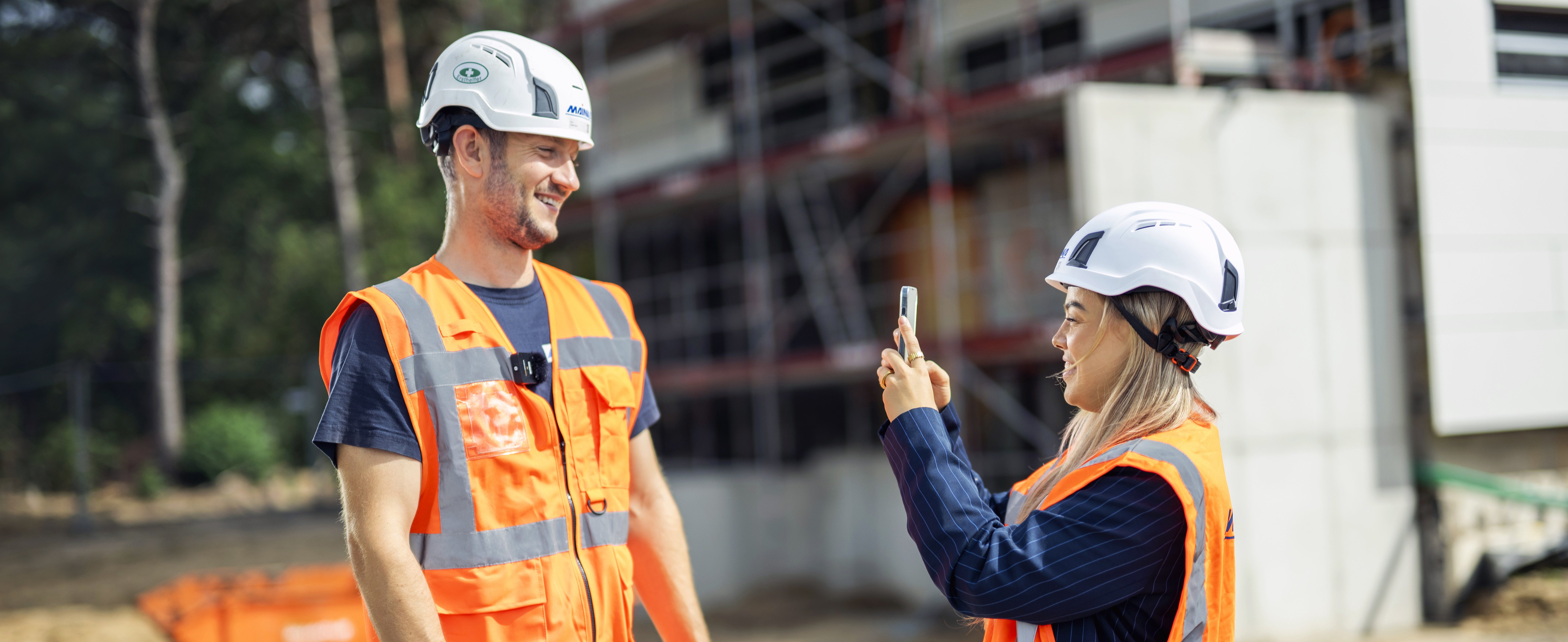 Zwei Bauarbeiter in orangefarbener Schutzkleidung, ein Mann und eine Frau, machen Fotos auf einer Baustelle.