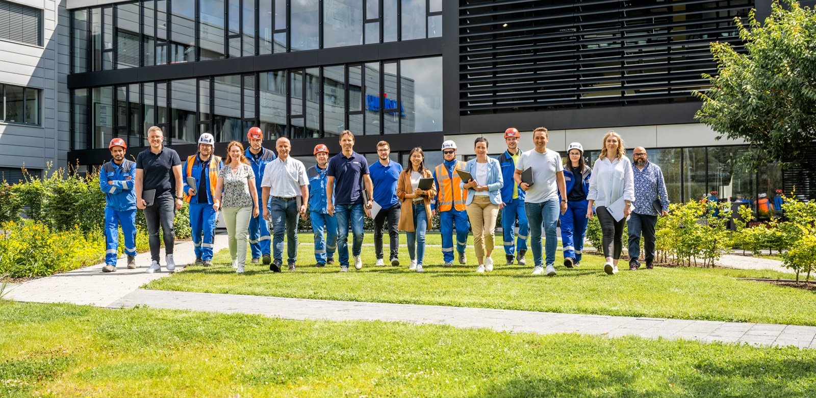 Gruppe von Menschen in Arbeitskleidung und Freizeitkleidung läuft auf einem Rasen vor einem modernen Gebäude.