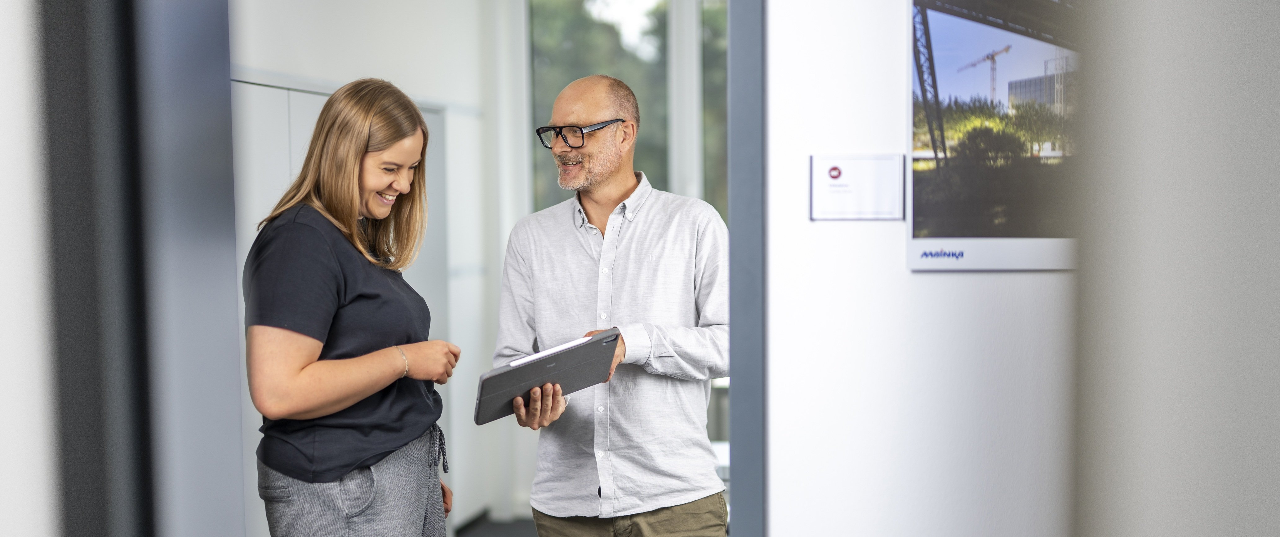 Zwei Personen stehen in einem Büroflur, eine hält ein Tablet, die andere schaut darauf.
