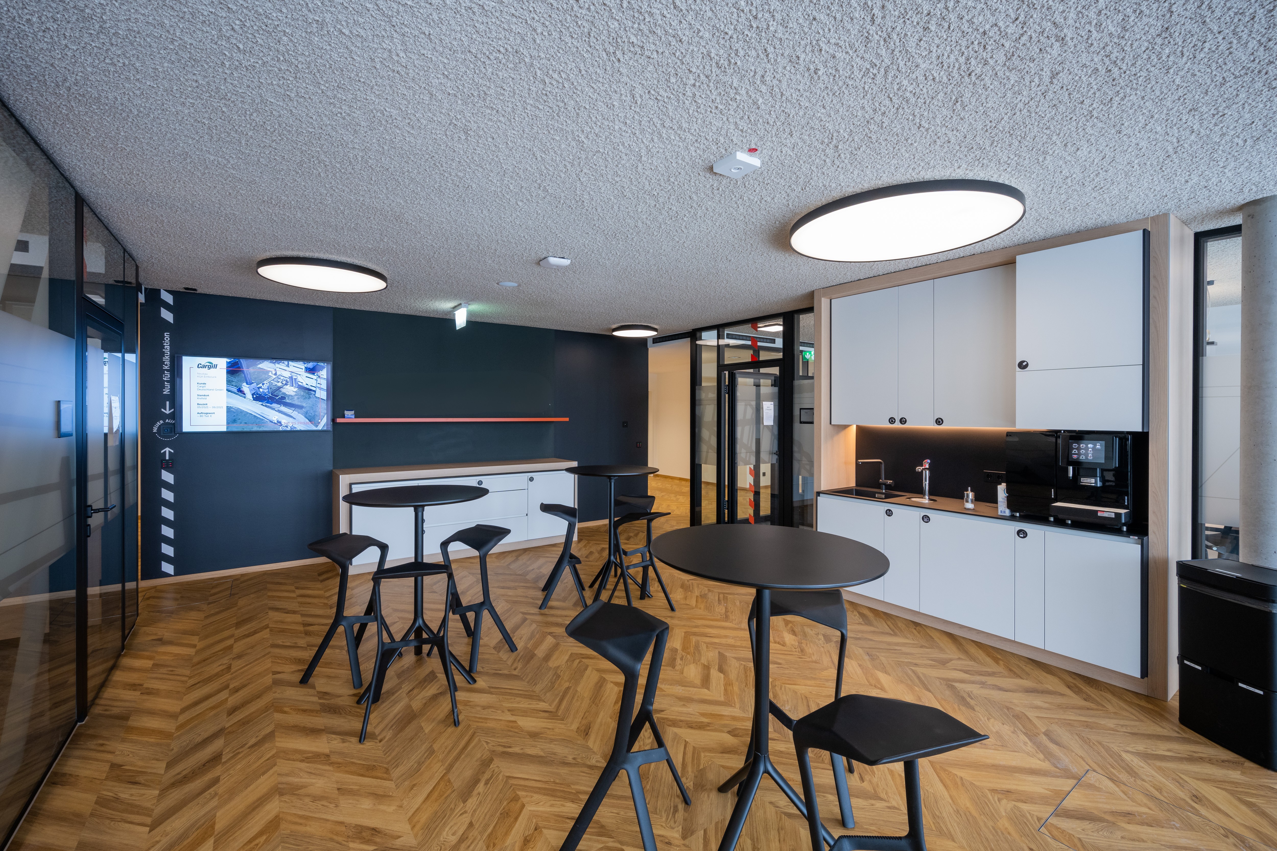 Moderne Teeküche mit schwarzen Tischen, Stühlen, Schränken, einem Waschbecken und einem Kaffeeautomaten.